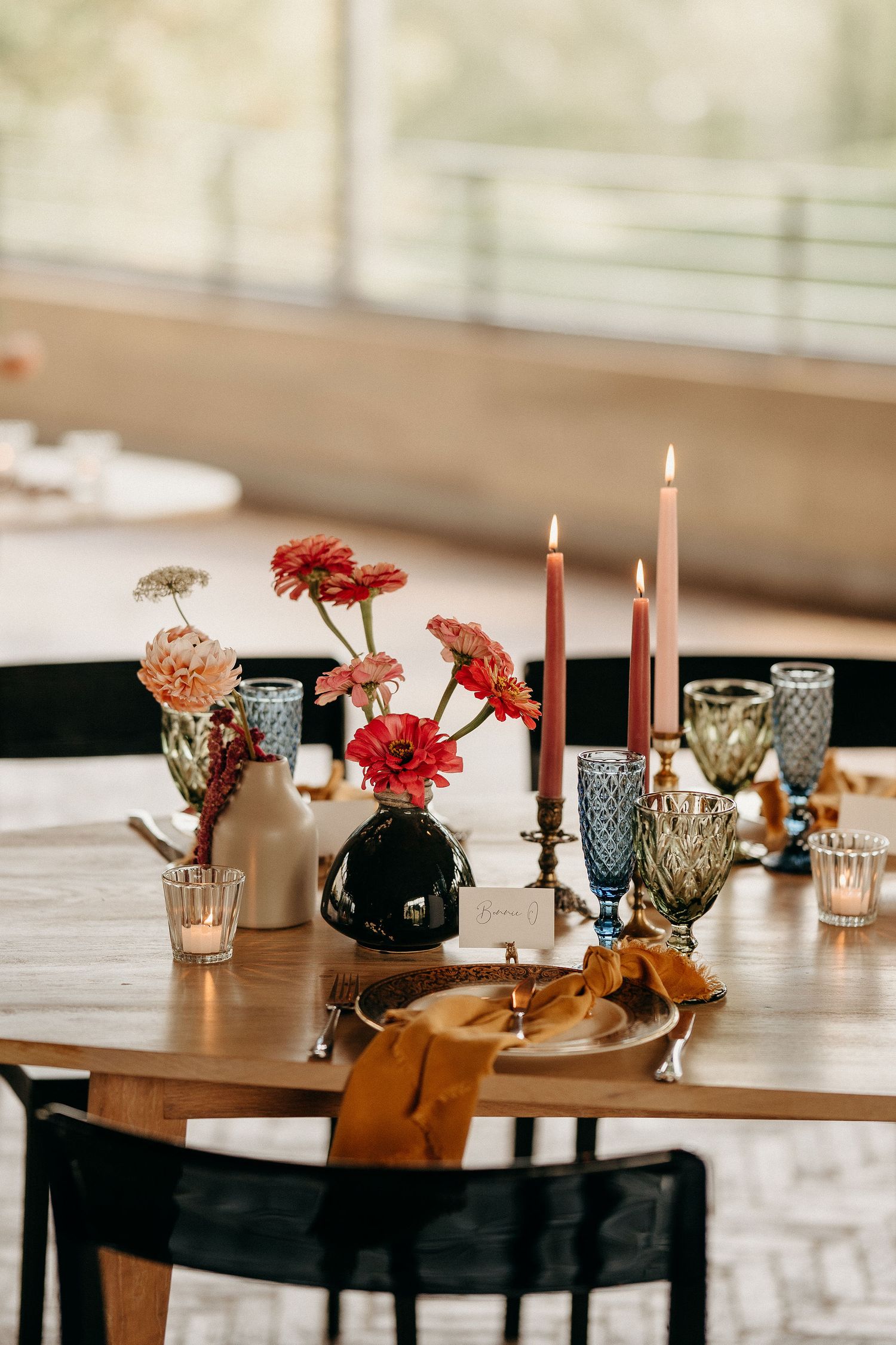 A romantic table setting with pink candles, colorful flowers in vases, and textured glassware on a wooden surface.