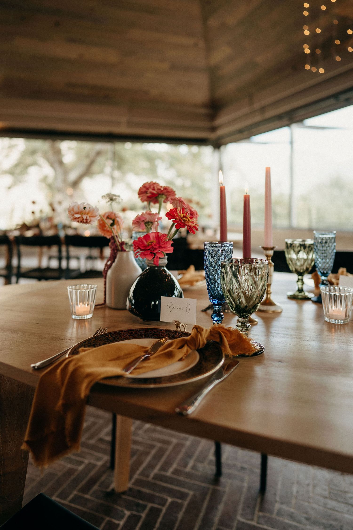 Elegant table setting with red carnations in vase, textured glassware and taper candles on wood surface under covered patio.