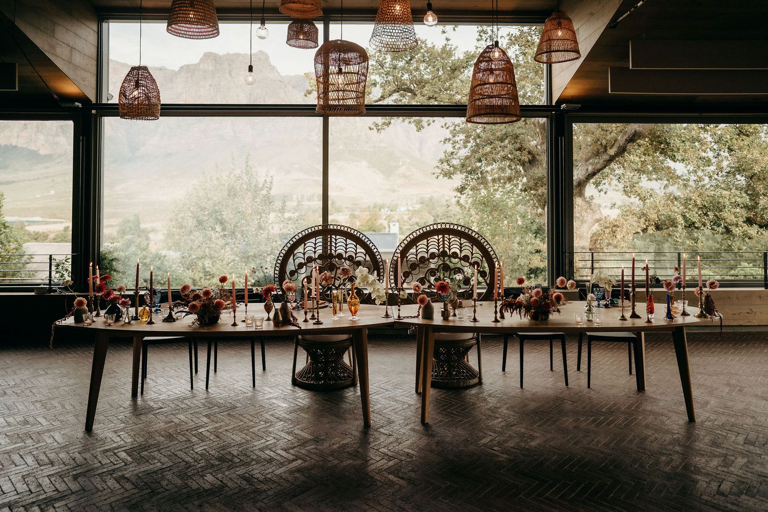 Long rustic dining table setup under vintage pendant lights with arched windows overlooking a natural landscape.
