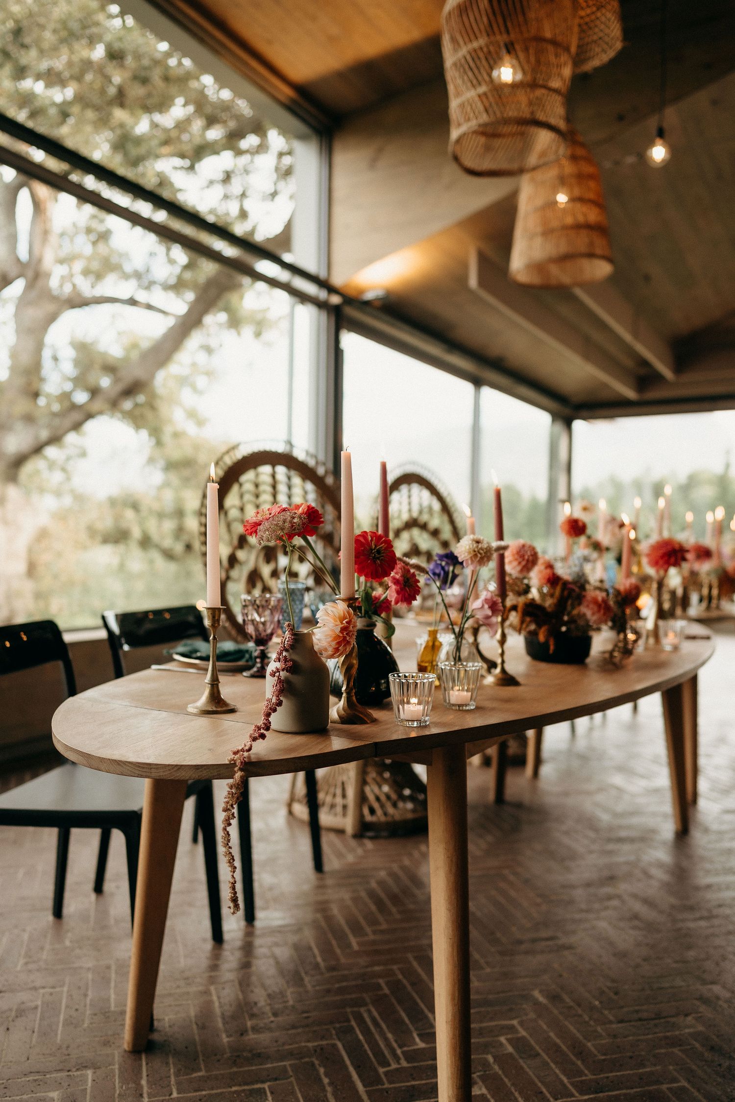 Elegant dining table decorated with red and pink floral arrangements and candlesticks under woven pendant lights by window.