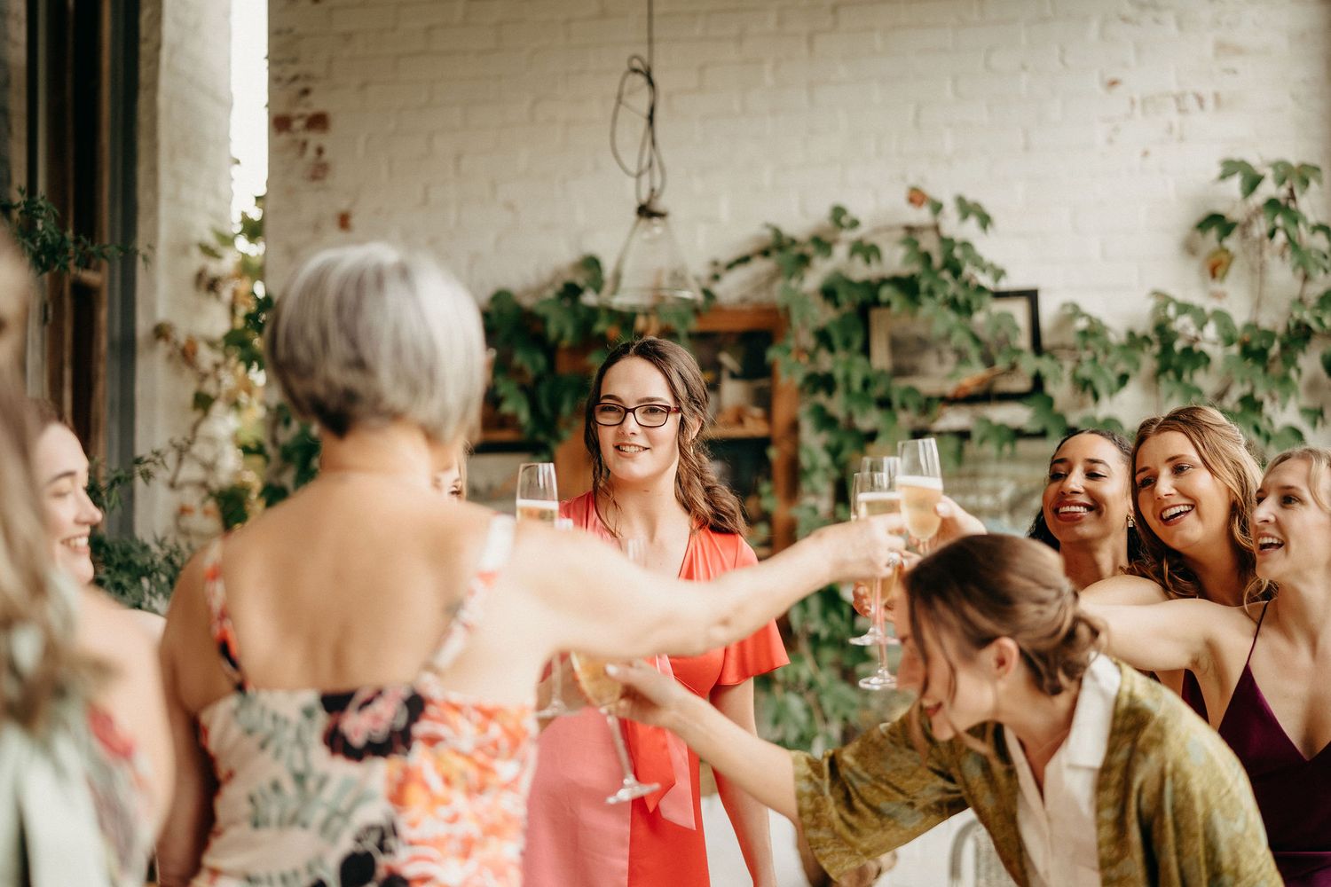 A group of friends raise their glasses in celebration at an indoor gathering with plants and brick walls.