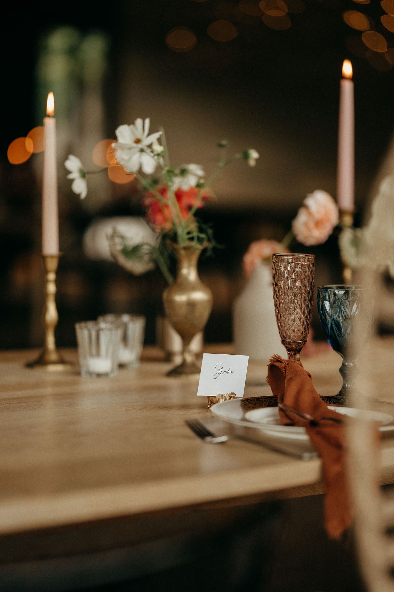 Elegant table setting with glowing taper candles, soft floral arrangements, and amber glassware on a wooden surface.