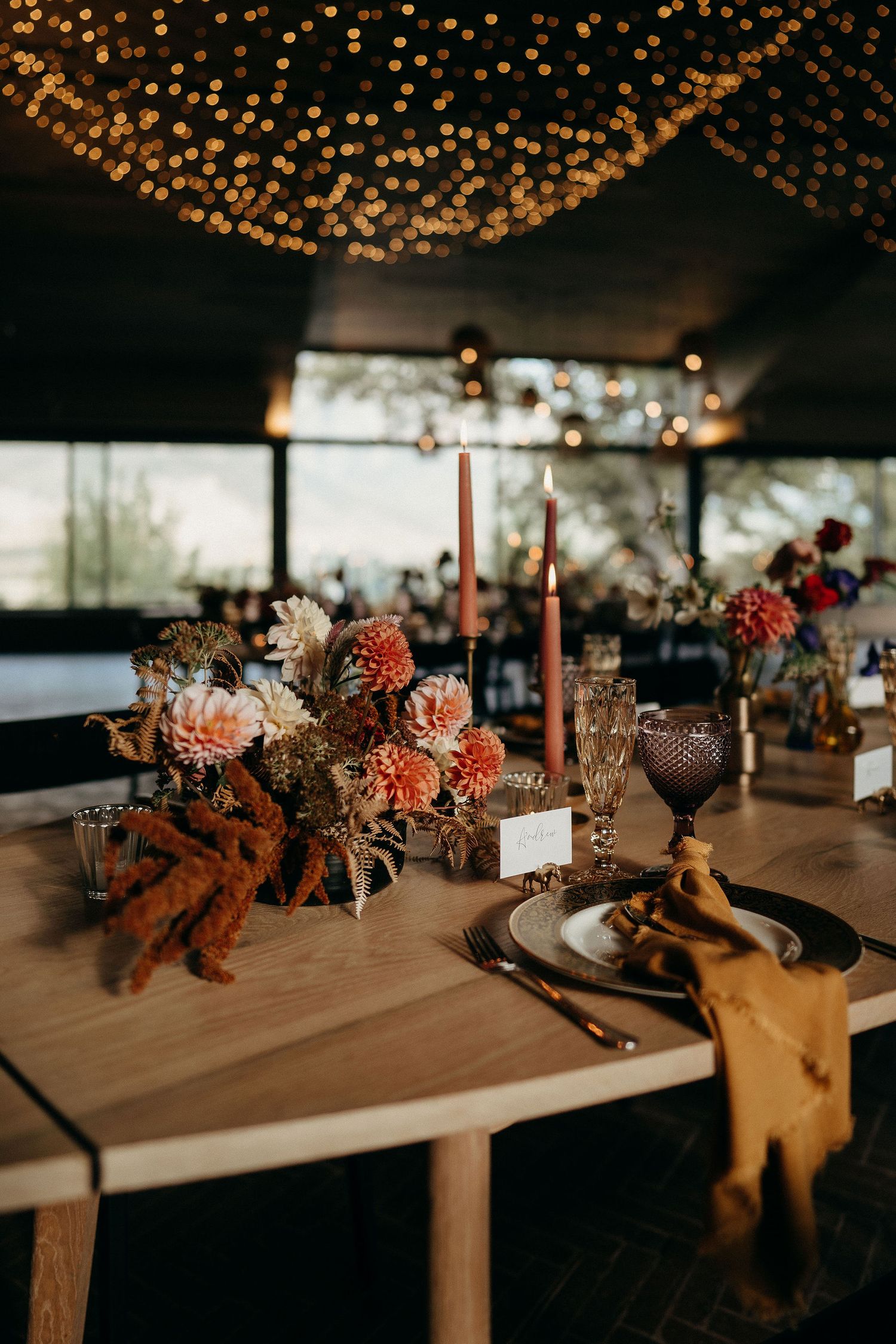 Romantic rustic wedding table setting with string lights, coral flowers, and vintage glassware on wooden surface.