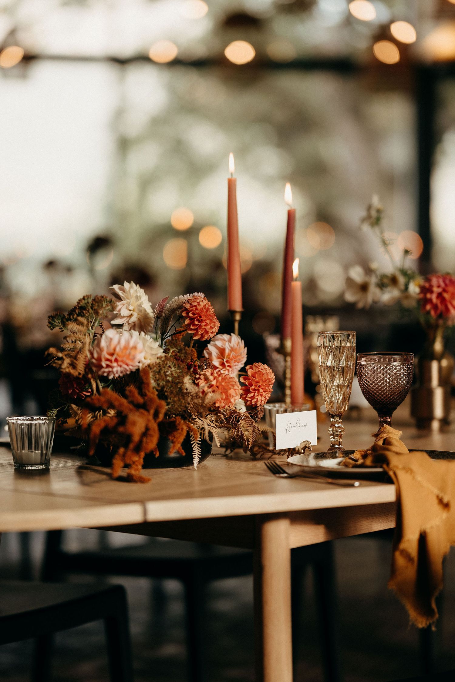 Moody autumn table setting with tall orange candles, vintage glassware and floral centerpiece in warm peachy tones.