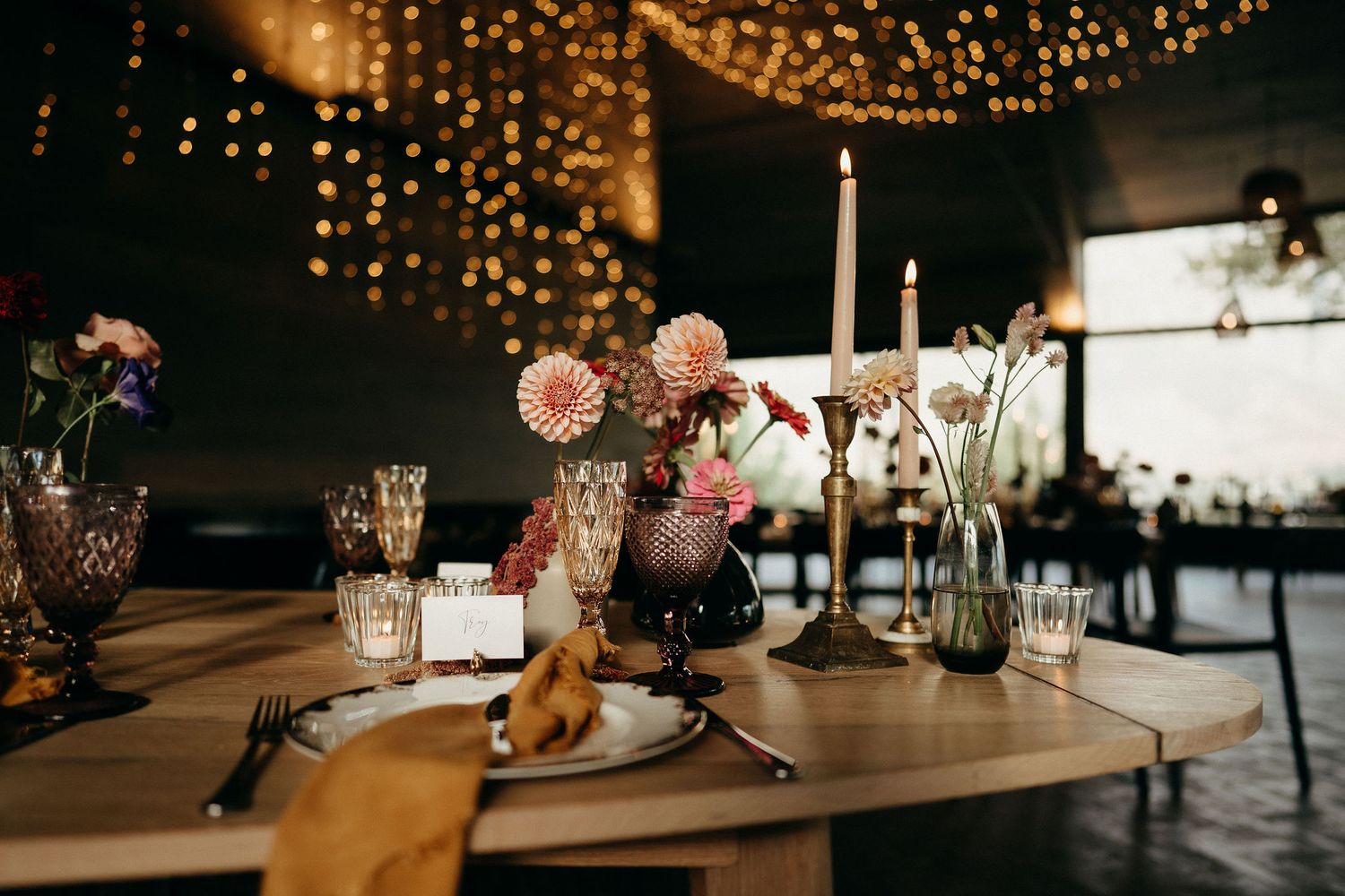 Elegant wedding table setting with string lights overhead, pink flowers, and tall white candles on rustic wooden surface.