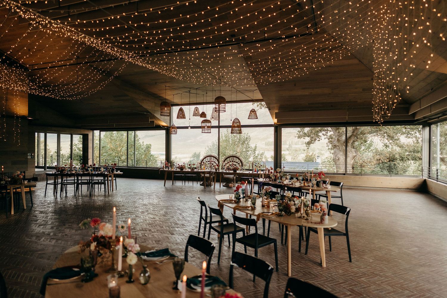 An elegant reception venue with string lights overhead, black chairs, and large windows overlooking a natural setting.