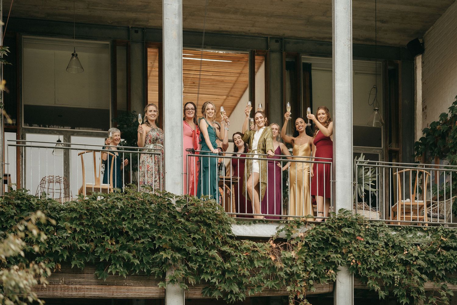 A group in colorful bridesmaids dresses stand together on an ivy-covered balcony during a wedding celebration.