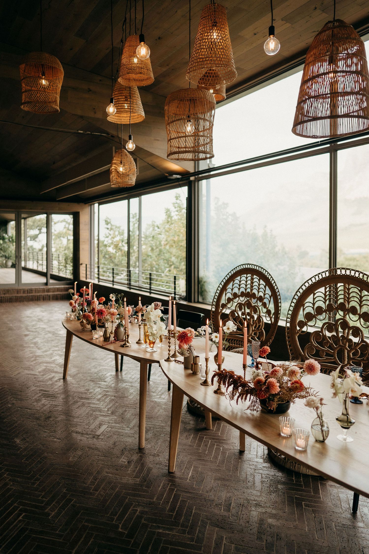 Long dining table with boho wicker lamps and peacock chairs set against floor-to-ceiling windows in a rustic venue.