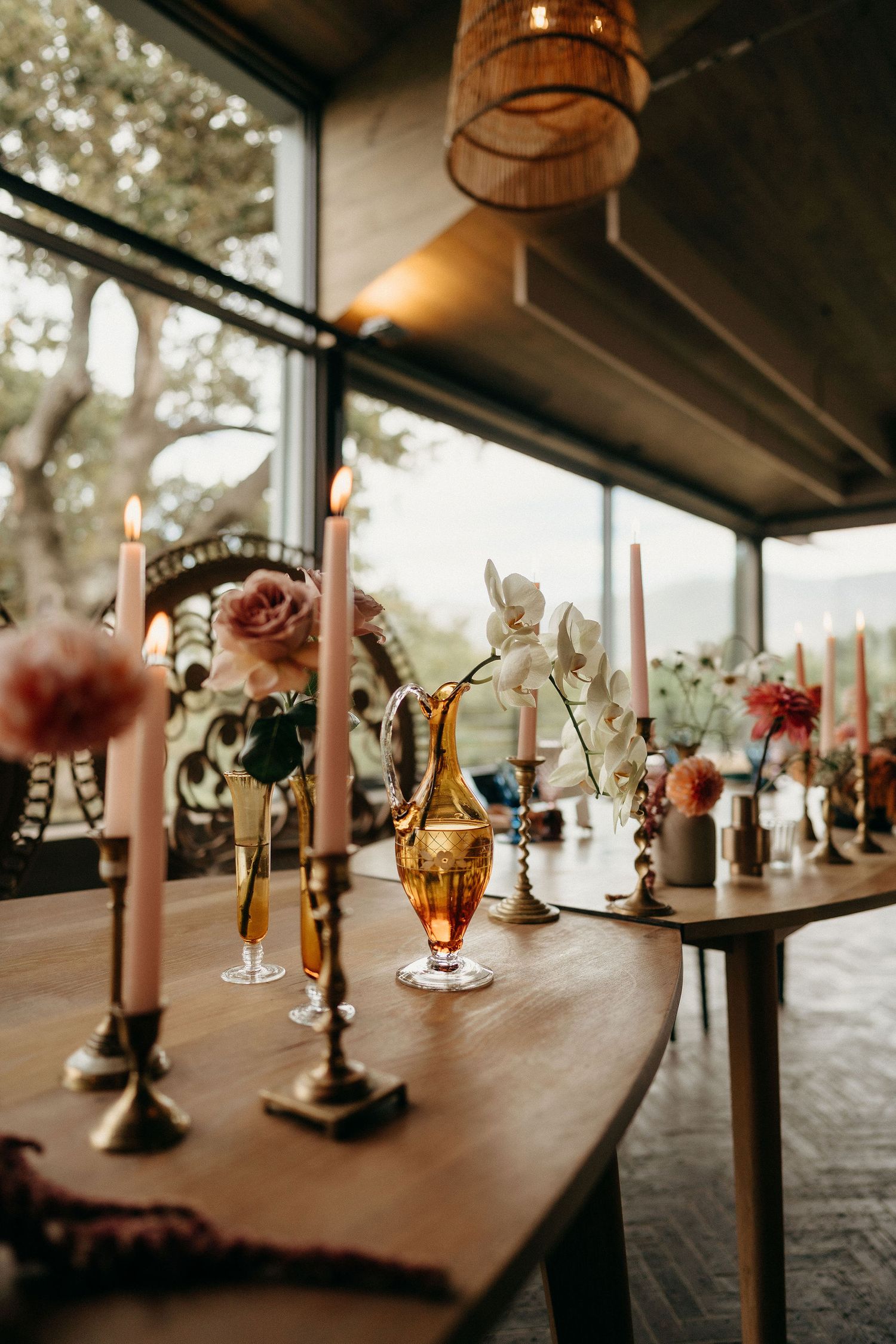 Rustic table decorated with amber glassware, taper candles, and floral arrangements in a sunlit room with pendant lighting.