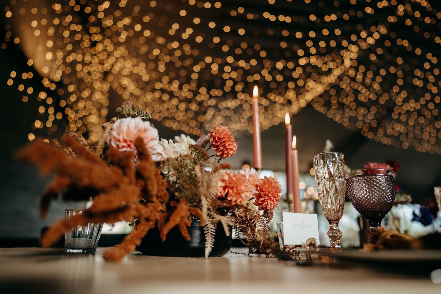 Elegant dinner table setup with glowing string lights, pink floral arrangements, and decorative rooster figurines.