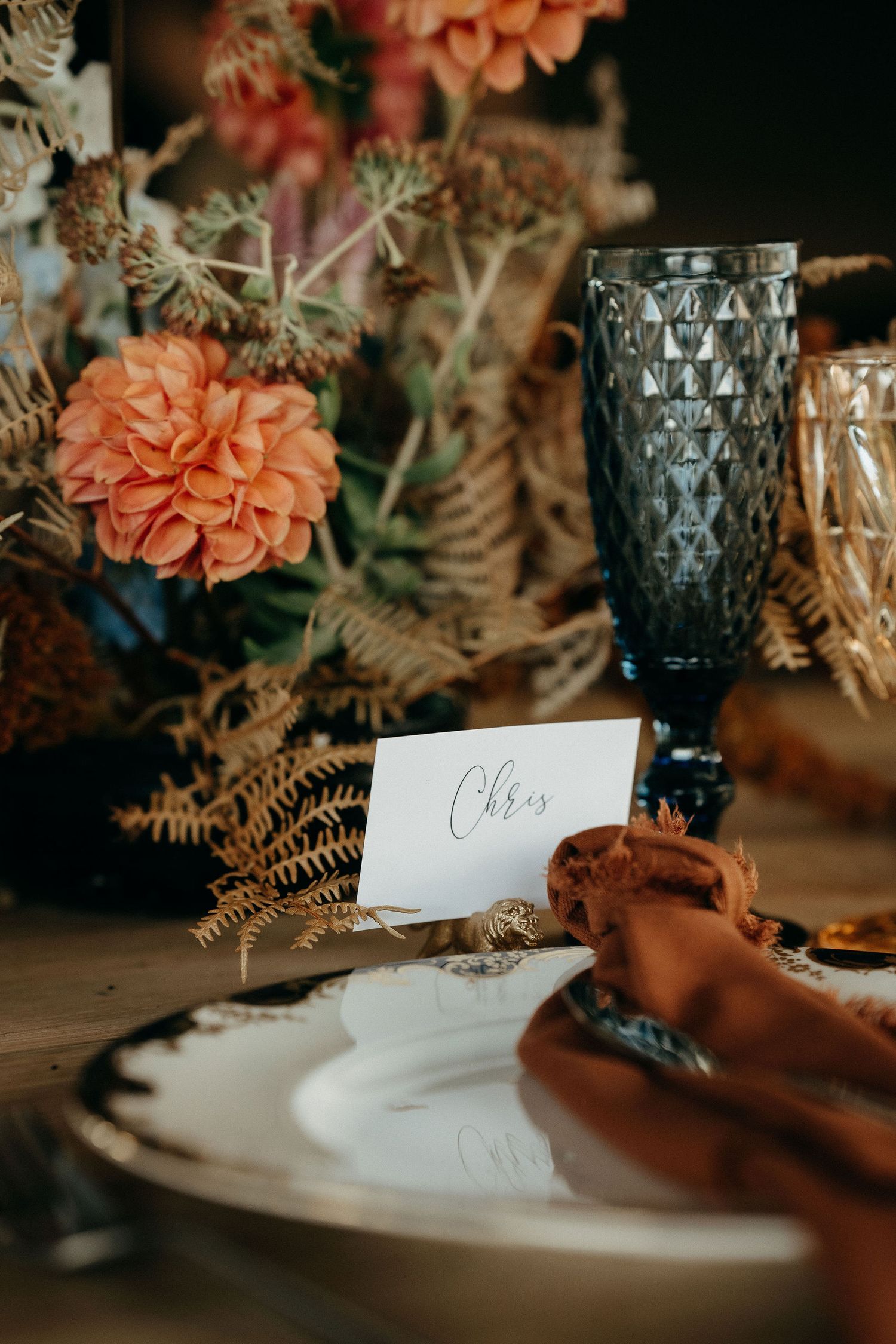 Elegant table setting with coral dahlias, dried ferns, blue glass goblet, and name card at romantic dinner.