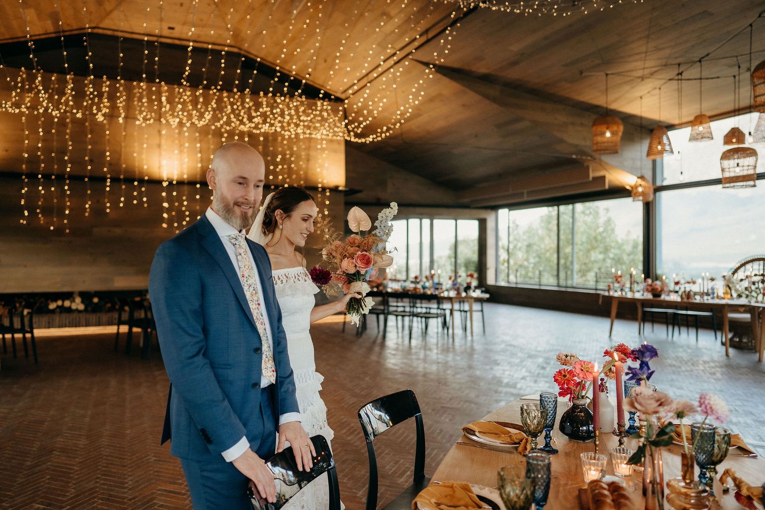 Couple celebrates at an elegant indoor venue with twinkling string lights and floral arrangements on wooden tables.