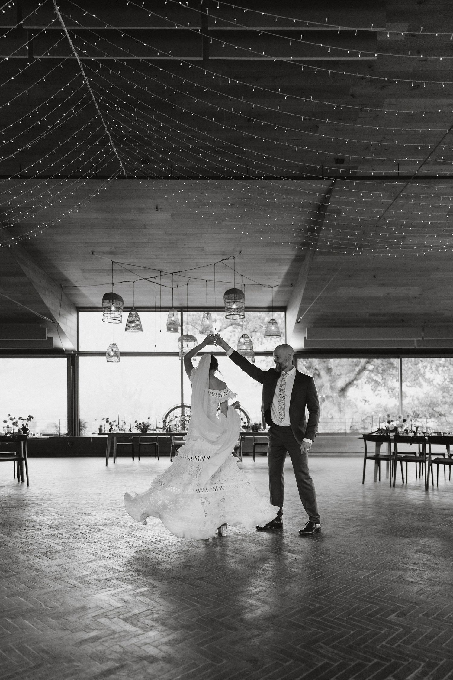 A black and white photo of a couple dancing in an empty reception venue with twinkling string lights overhead.