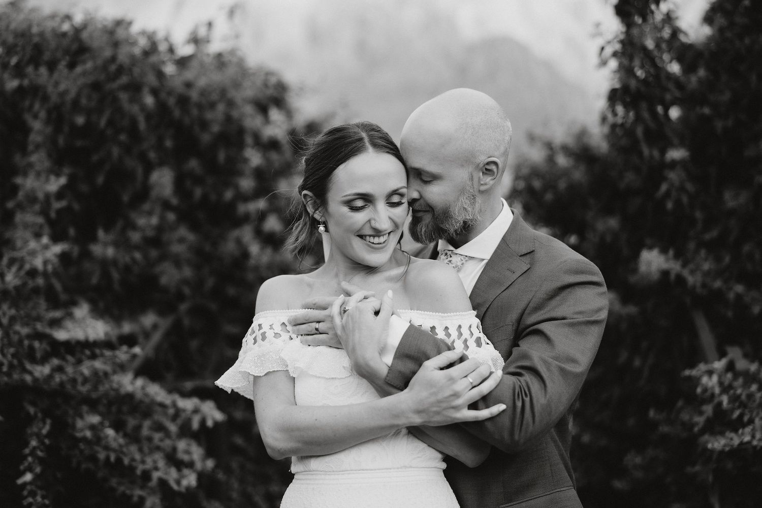 A tender black and white wedding photo of a couple embracing outdoors with mountains and foliage in the background.