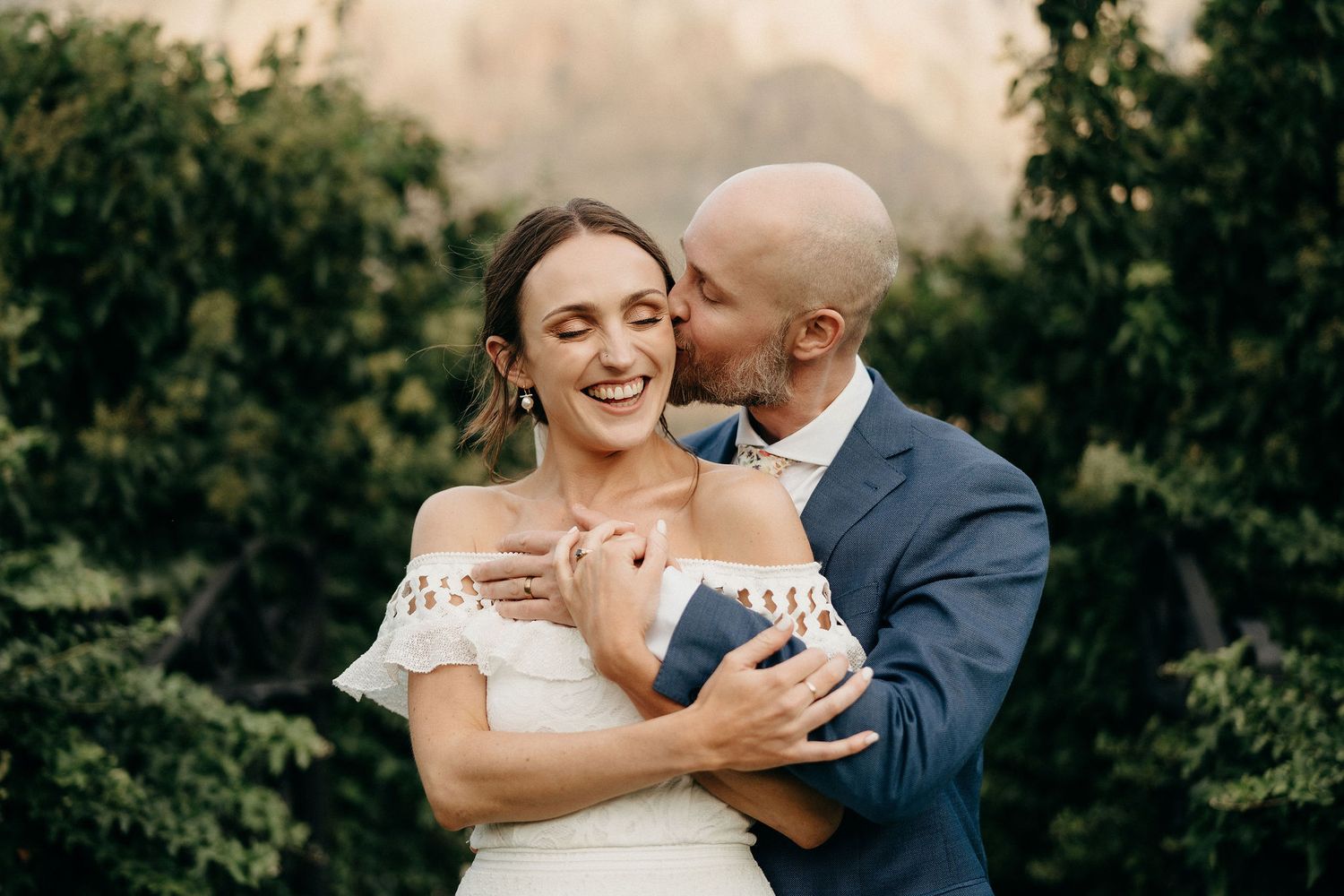 A joyful couple embraces outdoors during their garden wedding, the bride wearing an off-shoulder gown and the groom in navy.