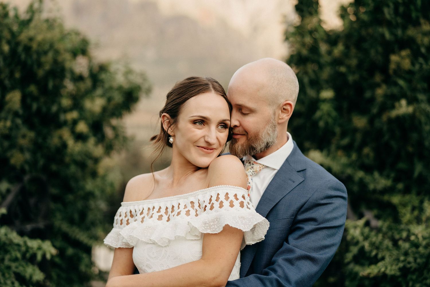 Romantic outdoor portrait of a couple embracing in a garden, with the bride wearing an off-shoulder lace dress.