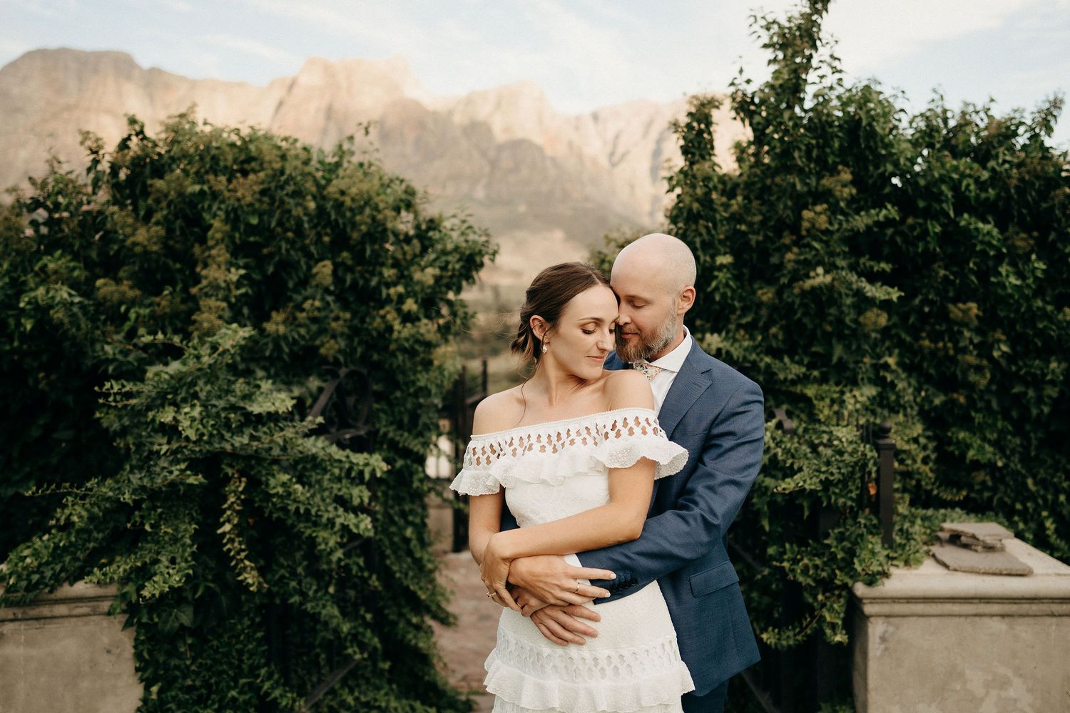 A couple embraces in a romantic garden setting with mountain scenery and lush greenery in the background.