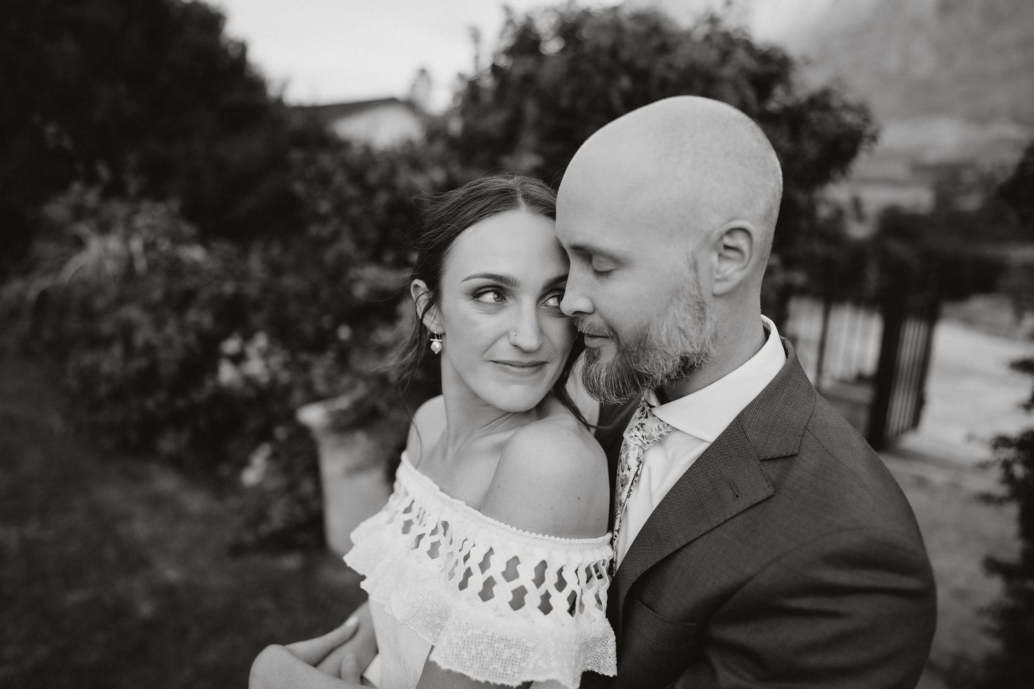 A black and white intimate portrait of a couple in wedding attire sharing a tender moment outdoors.