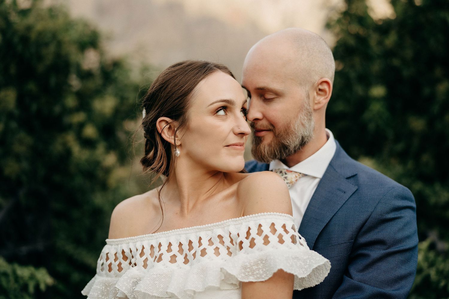 A romantic couple embraces during an outdoor wedding photoshoot wearing elegant attire against a green natural backdrop.