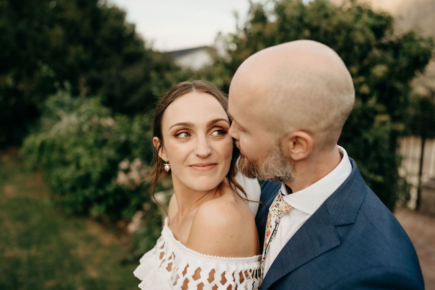 A romantic outdoor portrait of a couple in wedding attire sharing a tender moment against a green garden backdrop.