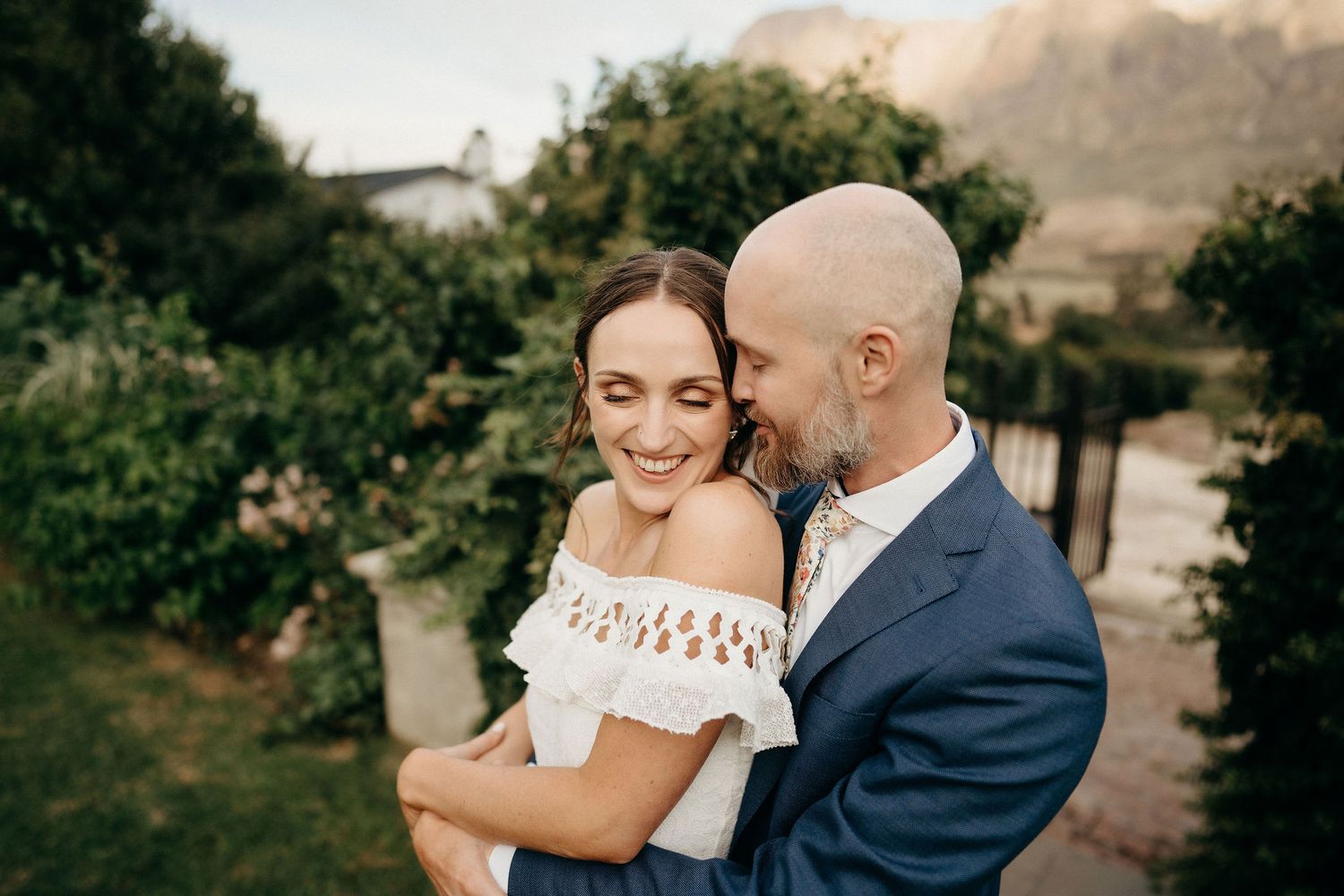 A romantic couple embraces outdoors during a wedding photoshoot with natural scenery in the background.