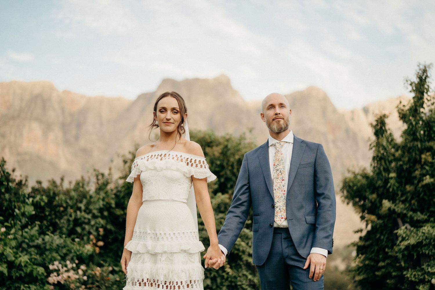 Two people hold hands at sunset with majestic mountain peaks and green foliage in the background.
