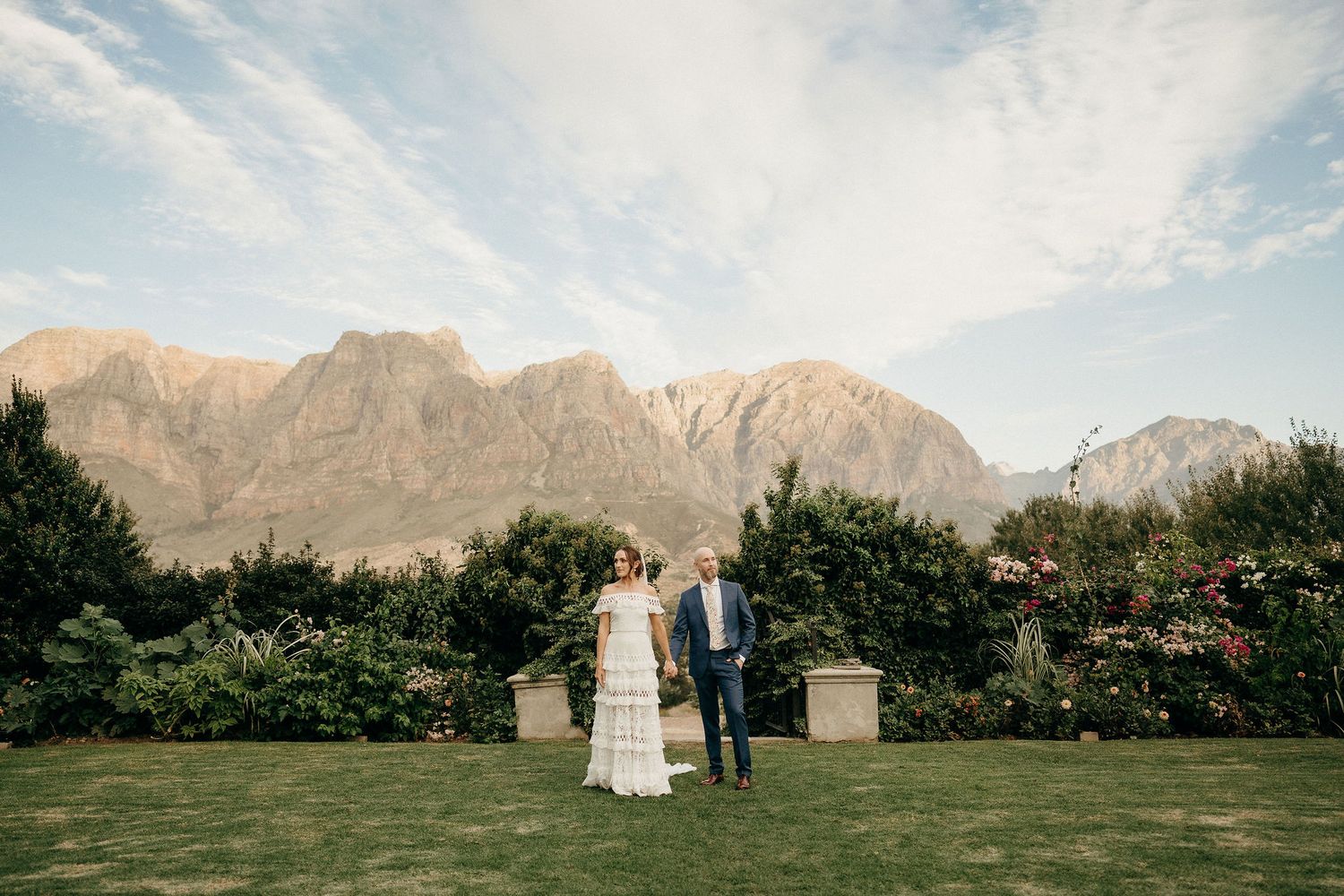 A couple stands on a lush green lawn with majestic mountain peaks and dramatic clouds forming a stunning backdrop.