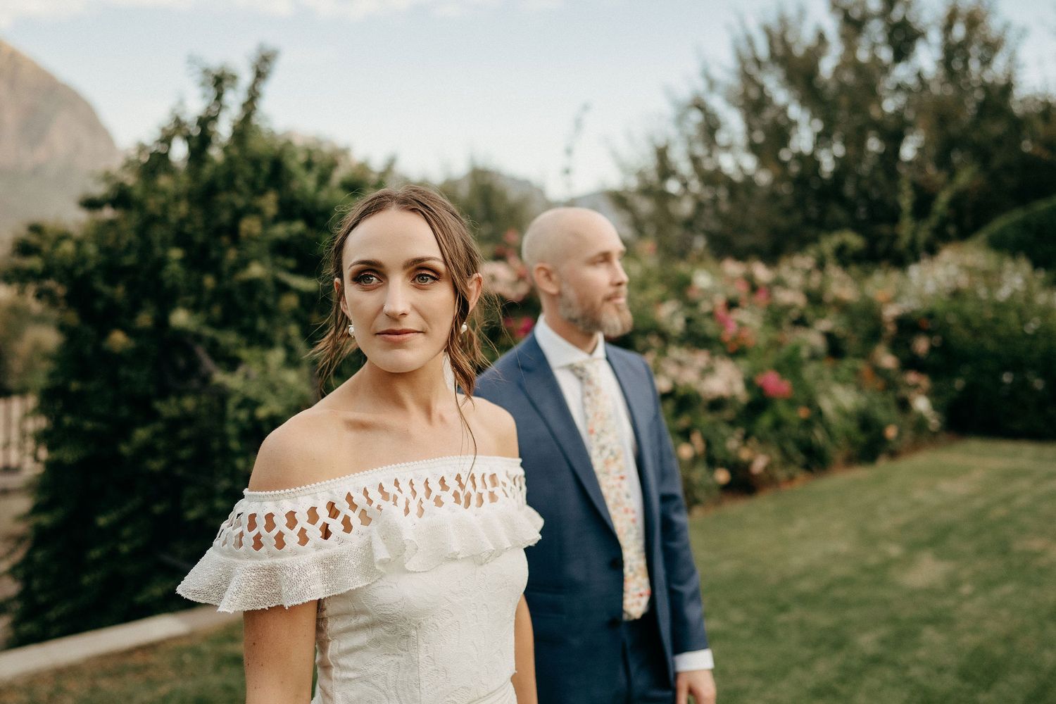 Couple in formal wedding attire pose in a garden setting with lush greenery and flowers in the background.