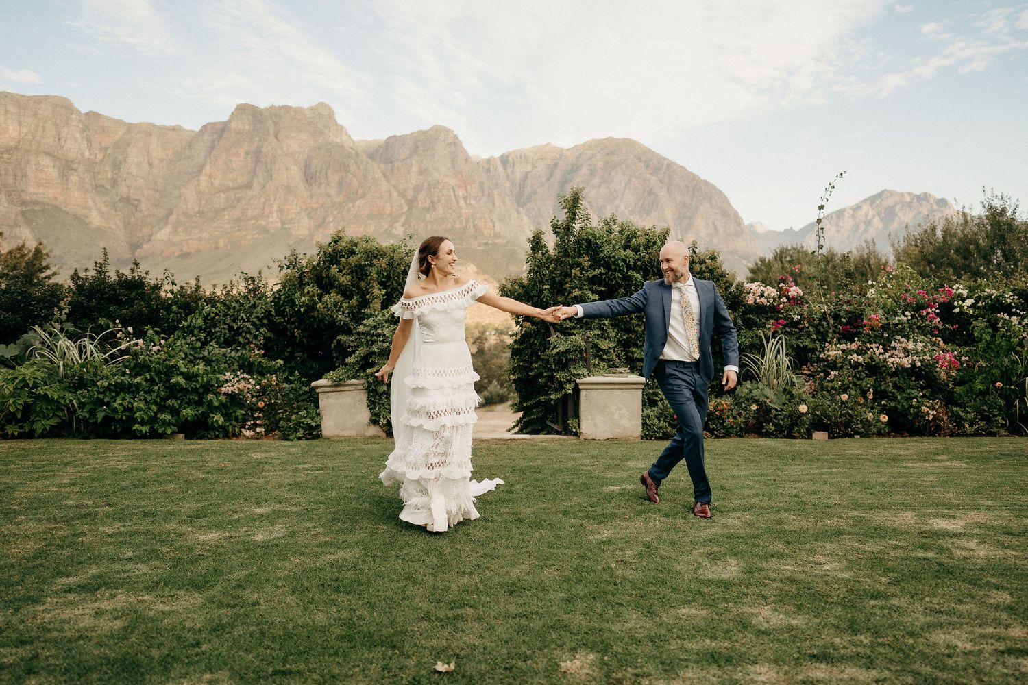 Two people dancing joyfully on a manicured lawn with dramatic mountains and gardens in the background.
