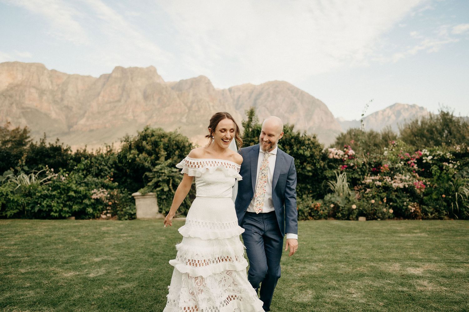 A couple joyfully running across a green lawn with majestic mountains and flowering bushes in the background.