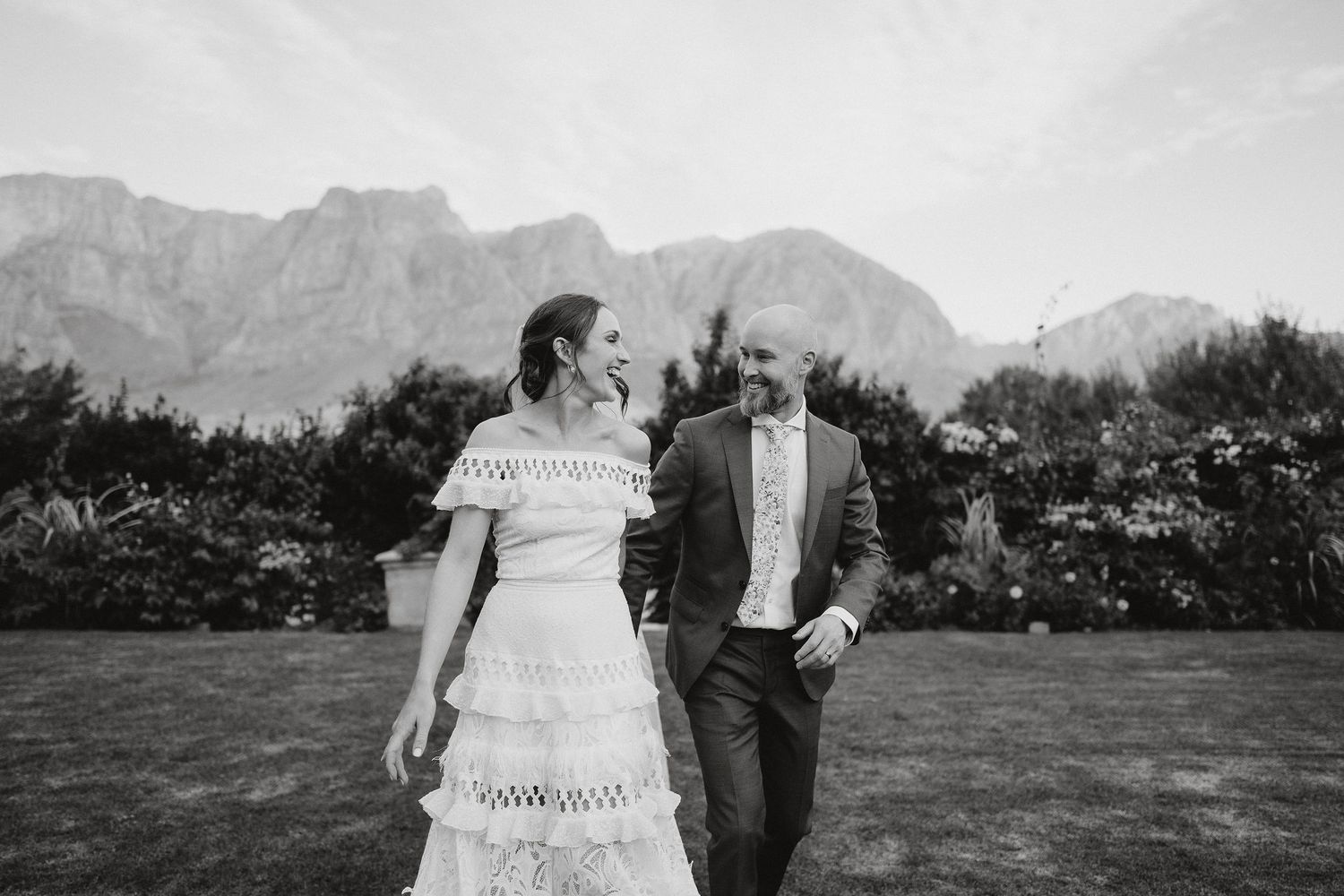 A couple walks together on grass with dramatic mountain peaks in the background in black and white.