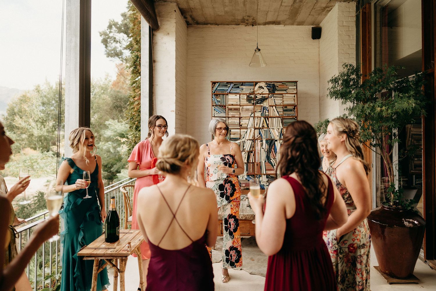 Wedding party in burgundy and emerald dresses gather on a covered porch during a warm afternoon celebration.