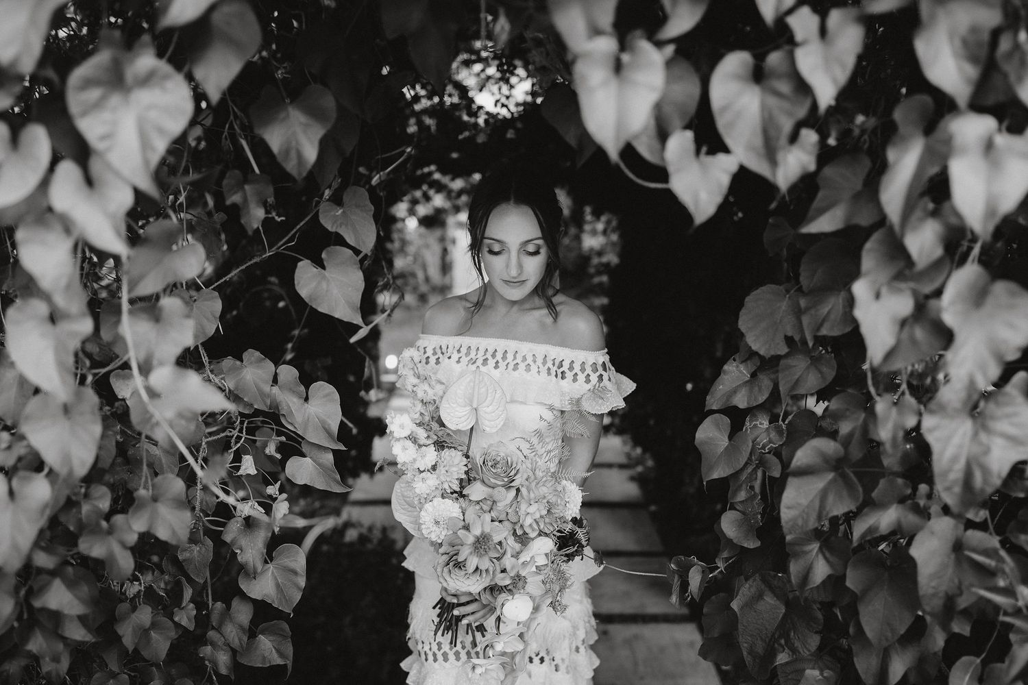 Black and white photo of bride in off-shoulder lace gown holding a luxurious white bouquet amongst leafy foliage.