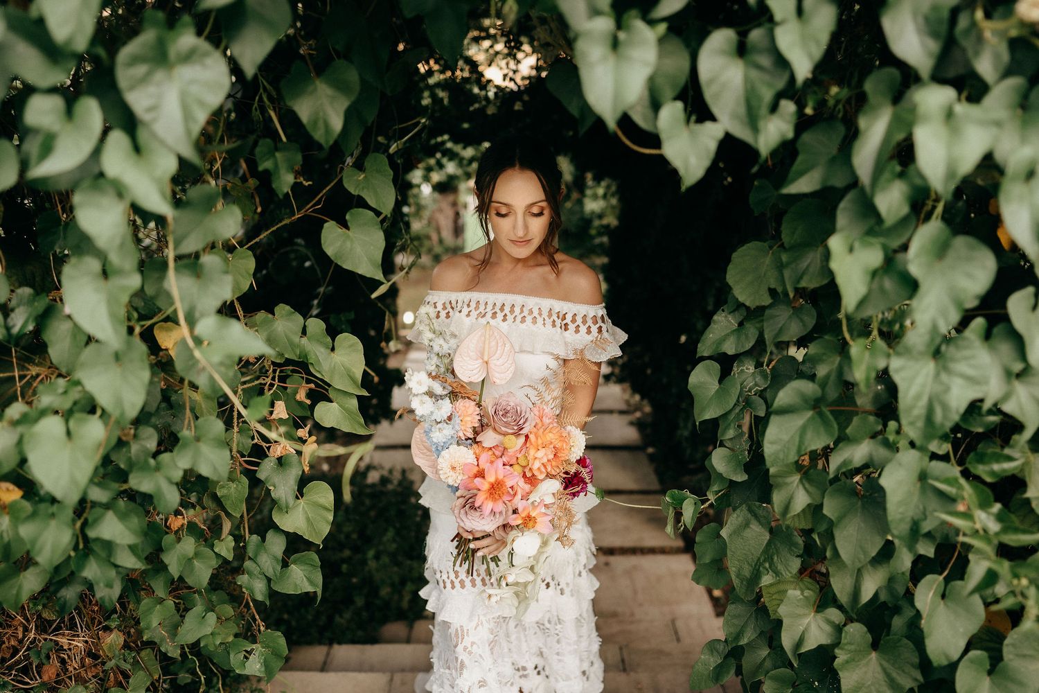 A bride in an off-shoulder lace wedding dress holds a peach and coral bouquet while standing amid lush green foliage.