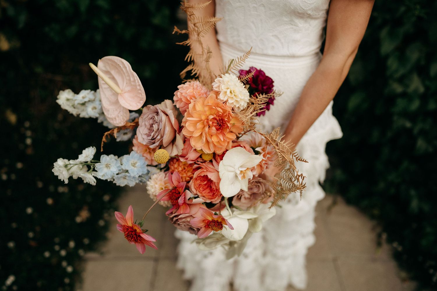 A bridal bouquet featuring coral, peach and burgundy flowers with dusty miller leaves against a white lace dress.