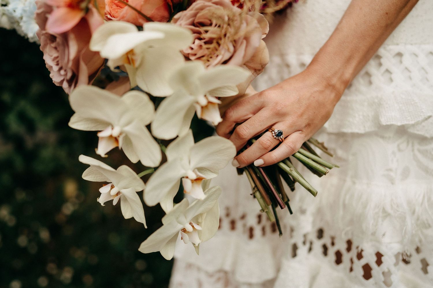Close-up of hands holding an elegant white orchid and rose bouquet against a lace wedding dress.