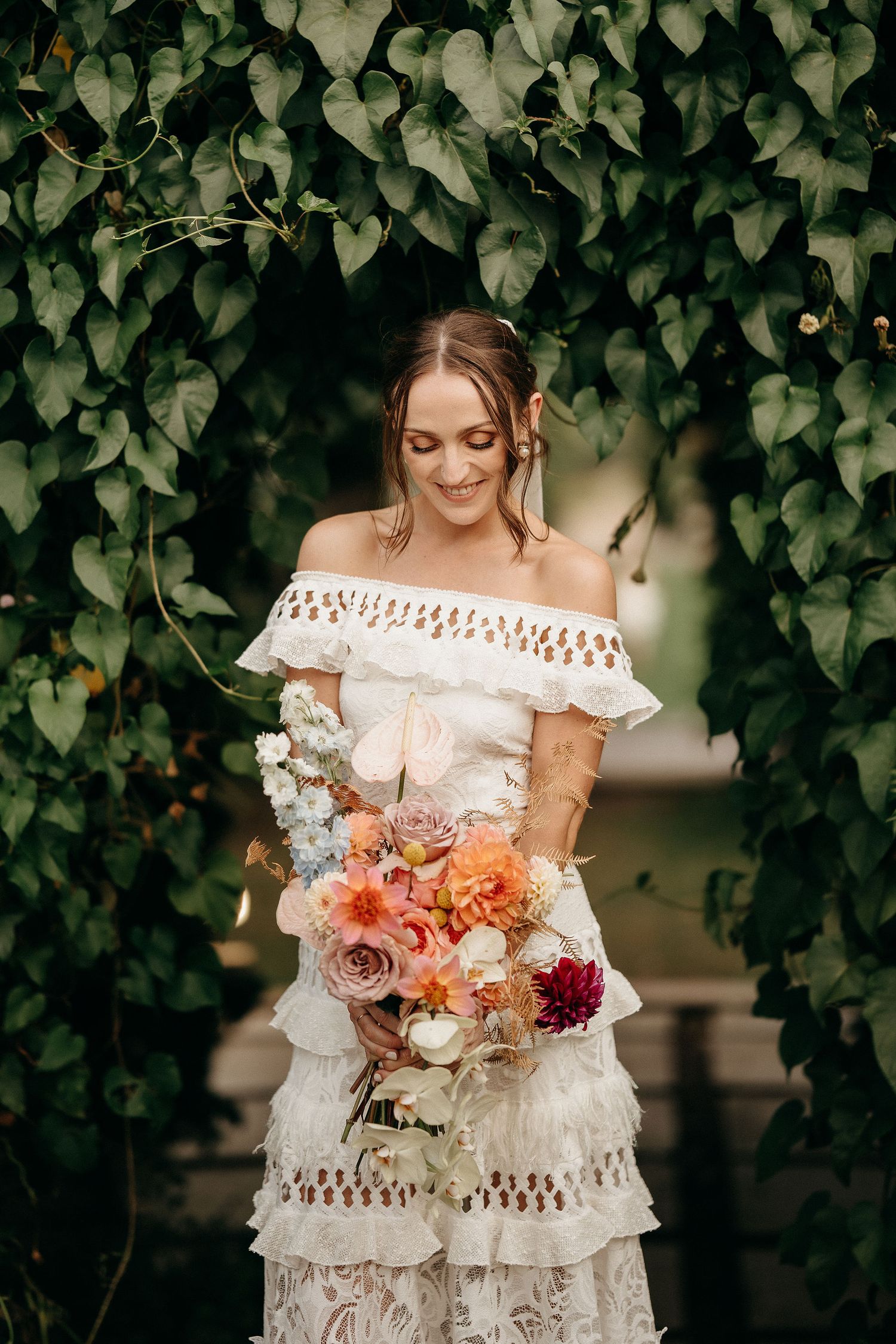 A lace off-shoulder wedding dress with bohemian details stands against a leafy backdrop, with a coral and blush bouquet.