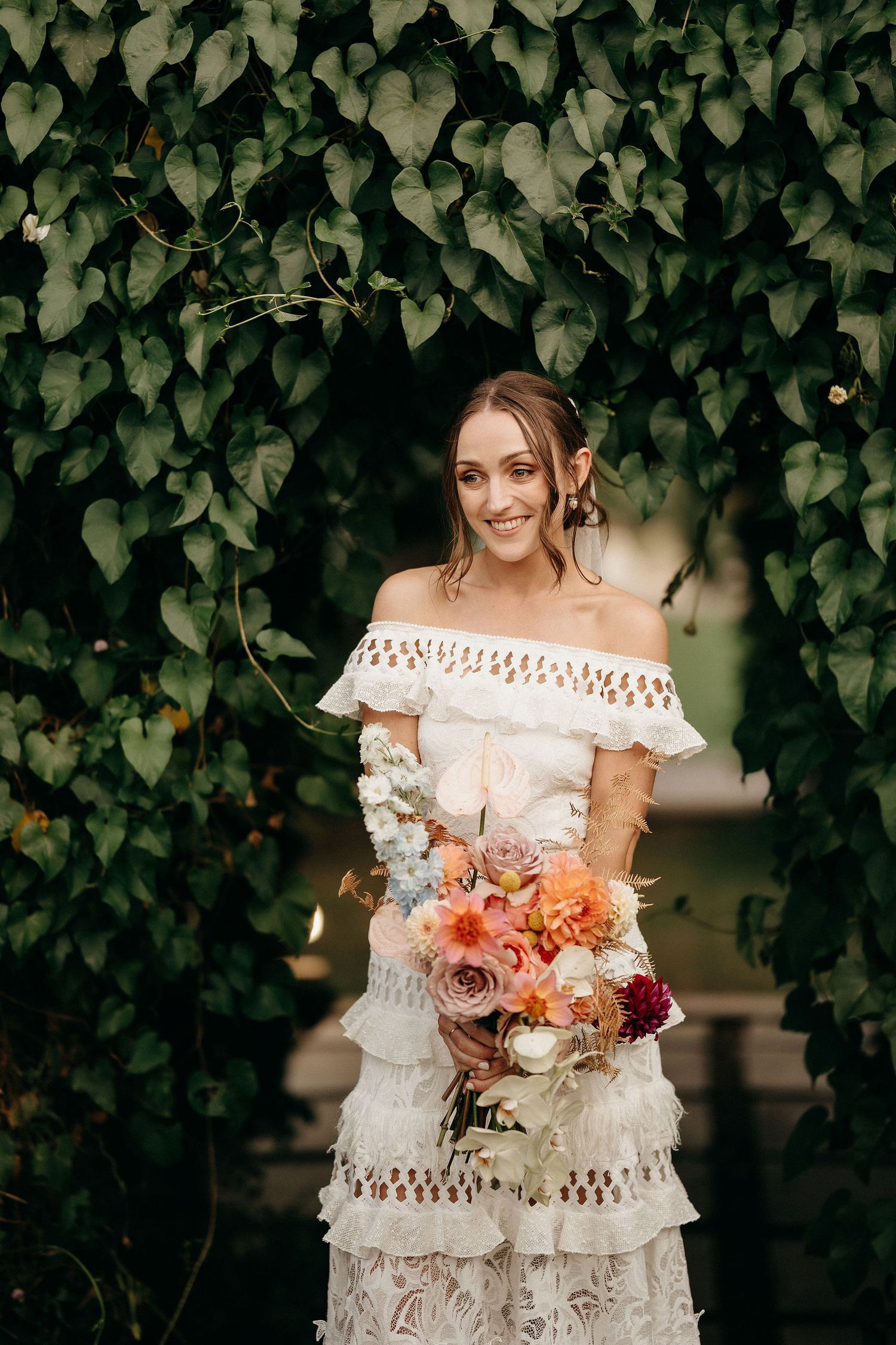A bride in an off-shoulder lace dress holds a coral and peach bouquet while standing against an ivy-covered wall.