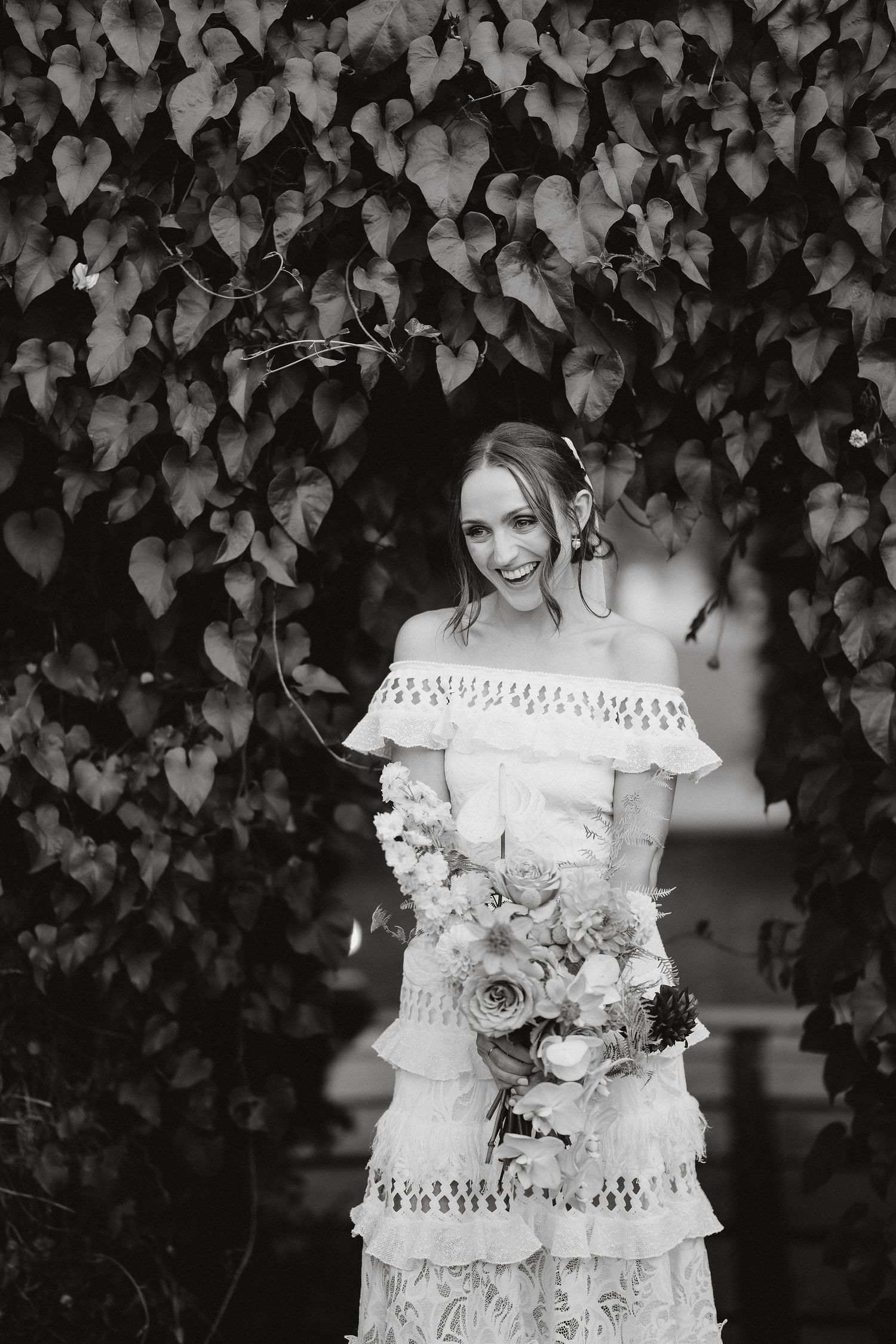A romantic black and white photo of a bridal look featuring a lace off-shoulder dress with tassels against an ivy wall backdrop.