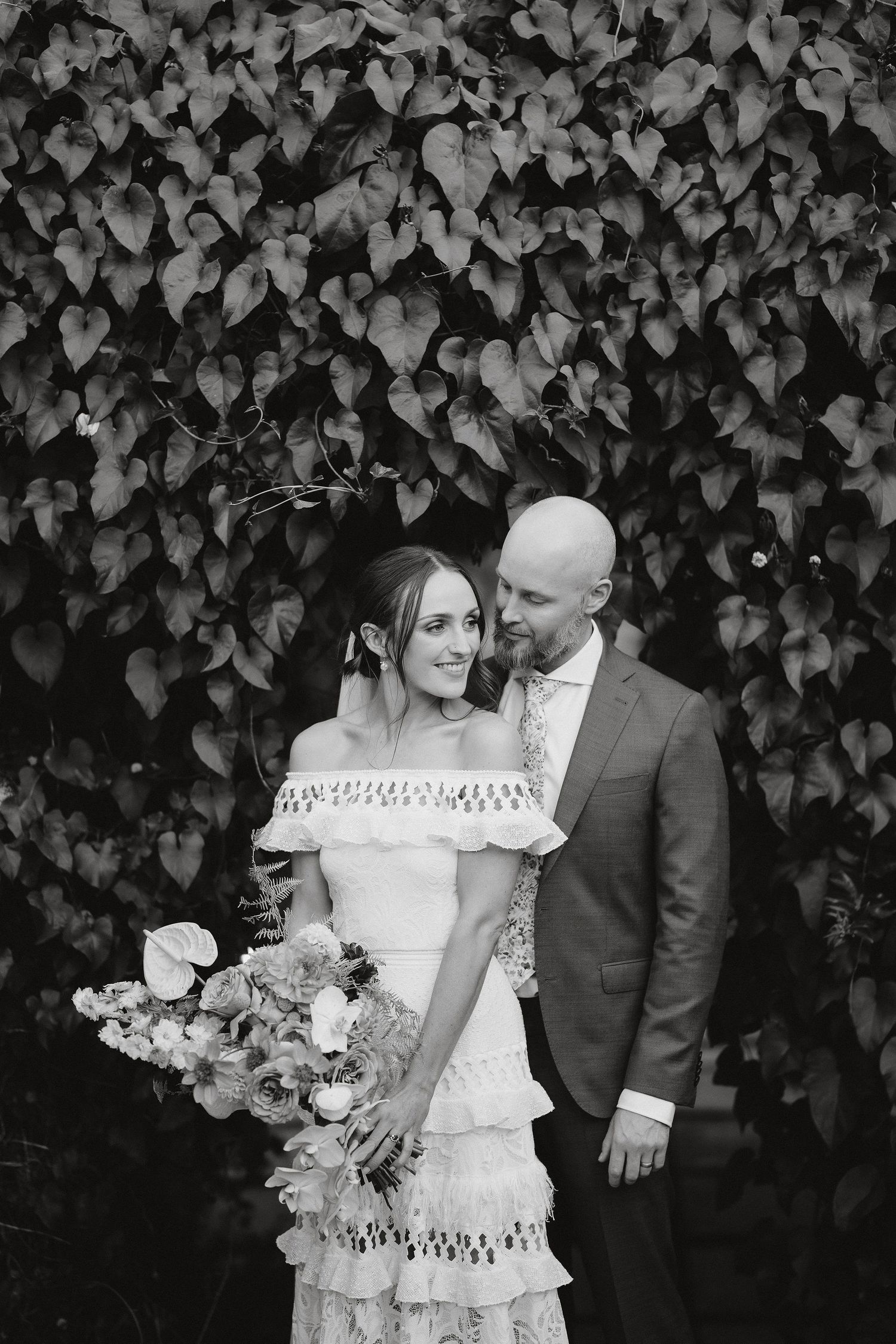 Black and white wedding portrait of couple in front of lush ivy wall with bride holding floral bouquet.
