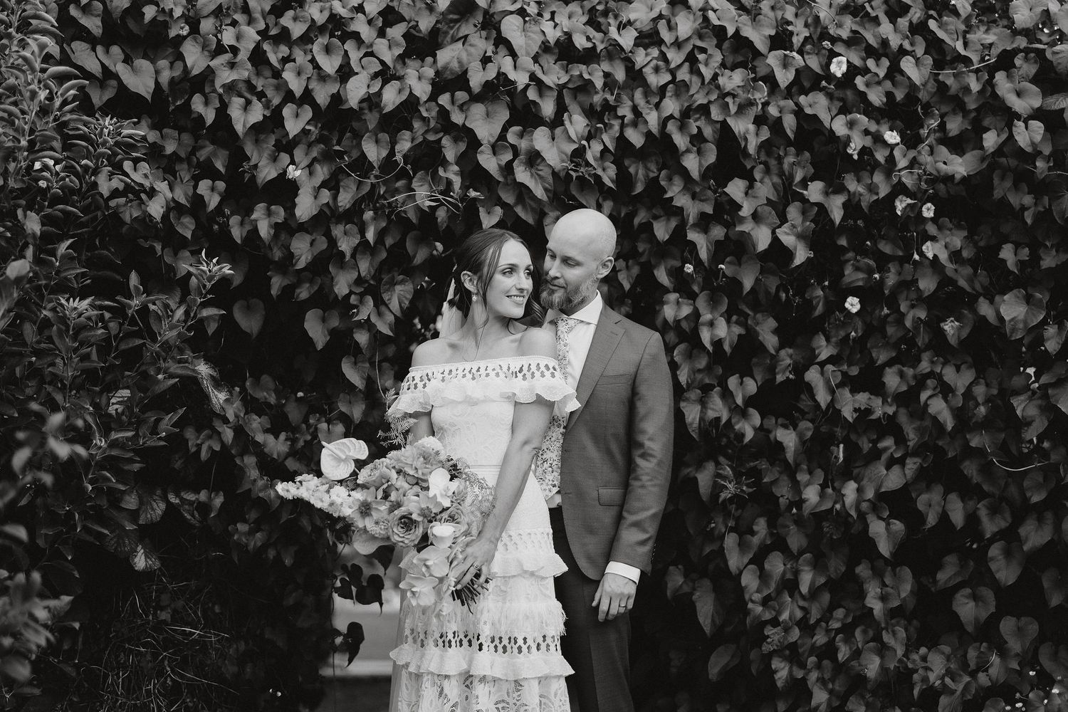 A couple poses together against an ivy-covered wall in a black and white wedding photograph.