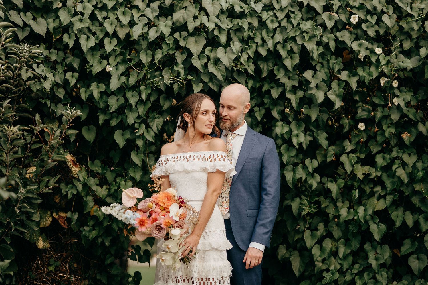 A couple poses against an ivy-covered wall while holding a colorful orange and pink wedding bouquet.