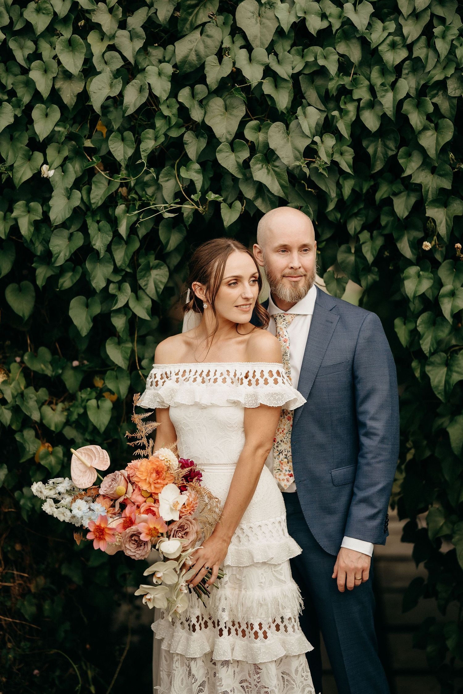 A couple poses in front of an ivy wall, wearing a tiered lace wedding dress and gray suit with a peach floral bouquet.