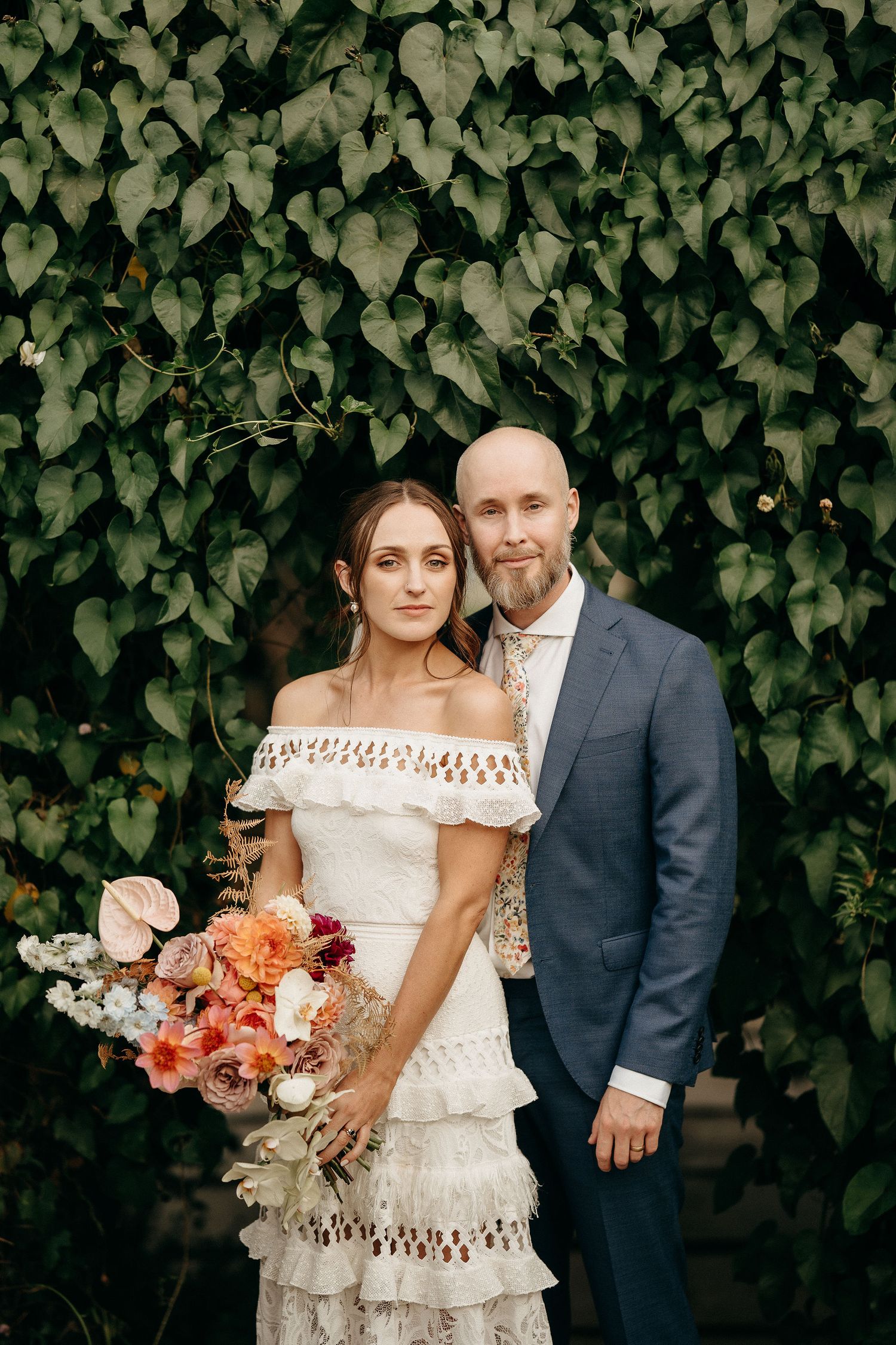 A wedding portrait against a lush green ivy wall backdrop with coral and white floral bouquet.