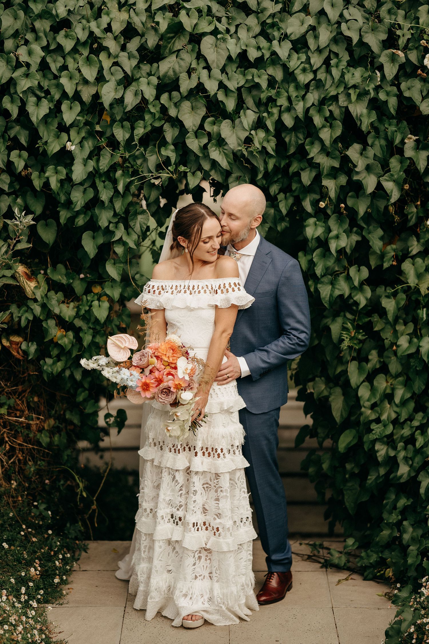 A couple embraces in front of an ivy-covered wall, with the bride wearing a bohemian lace dress and holding coral flowers.