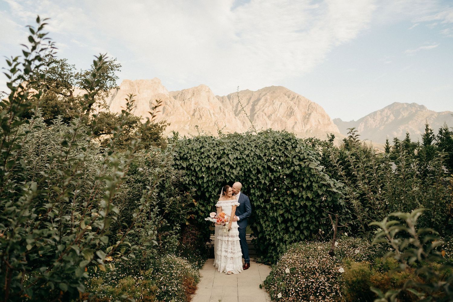 A couple stands embracing on a garden path with dramatic mountain peaks rising in the background at sunset.