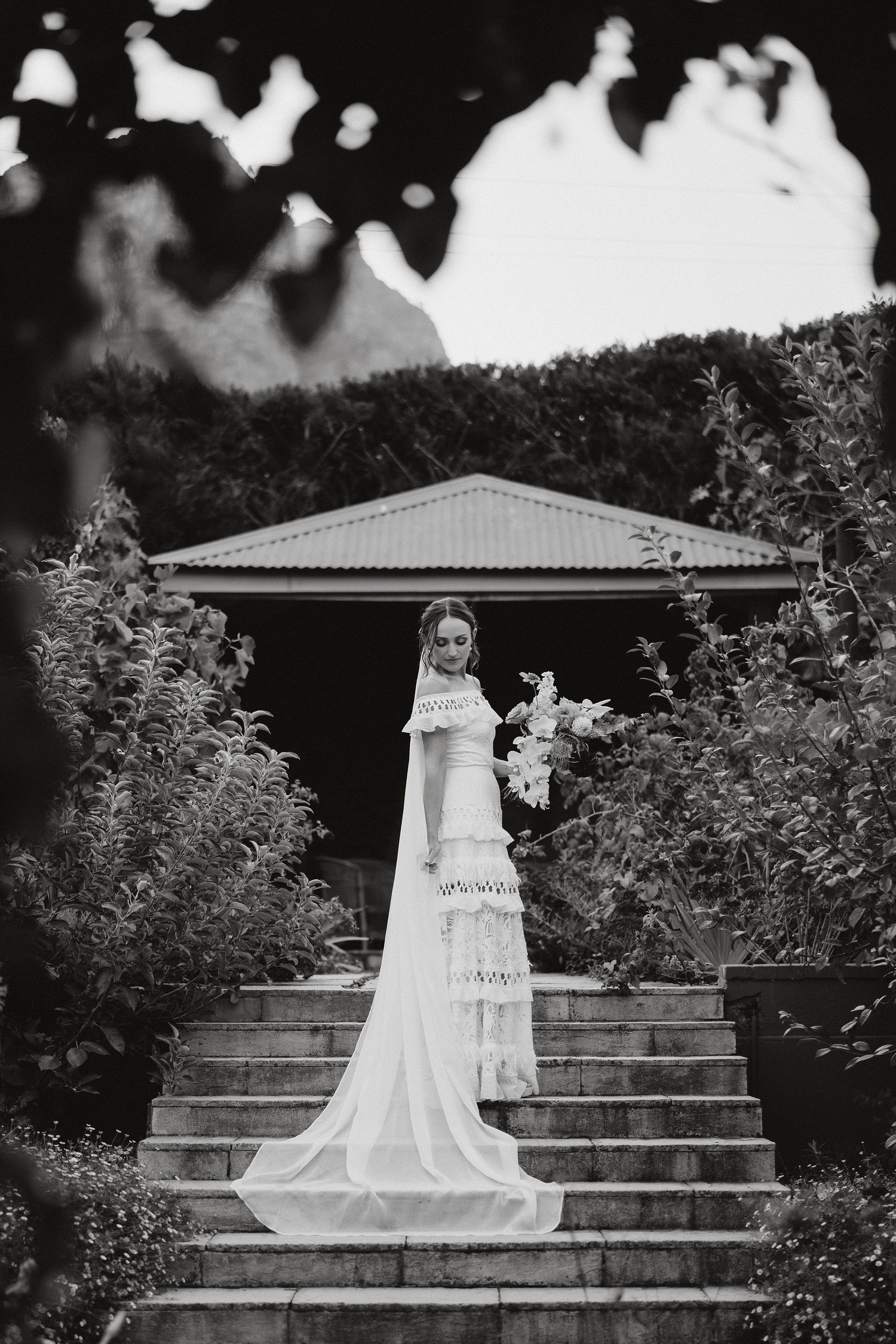 Black and white artistic wedding photo showing person in elegant white gown and veil standing on outdoor stone steps.