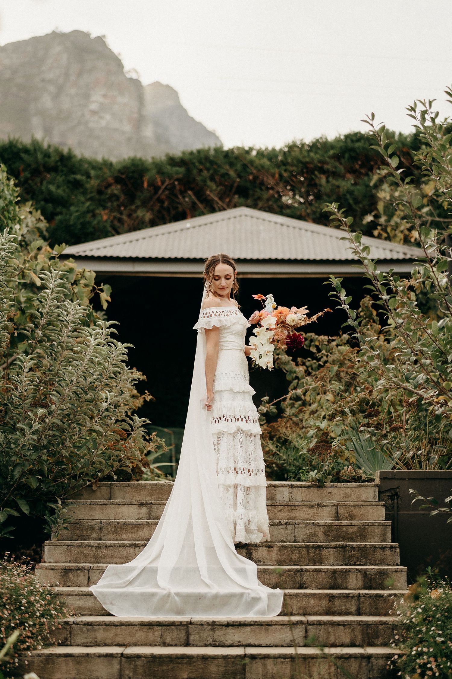 Elegant lace wedding dress photographed on garden stairs with mountain backdrop and rustic building in background.