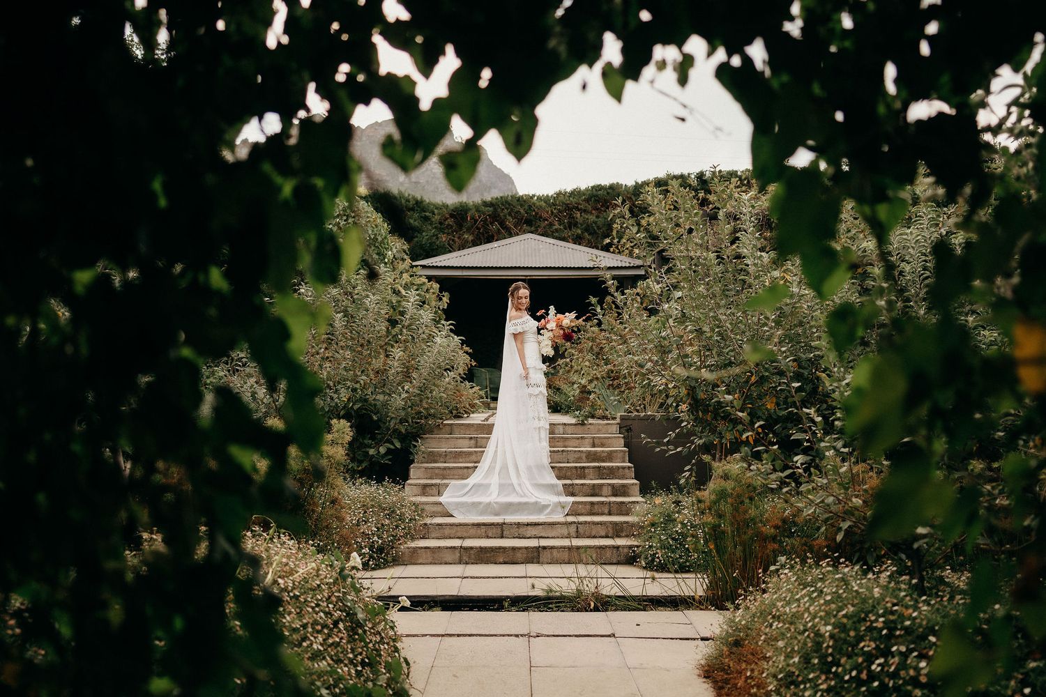 A person in a flowing white wedding dress stands beneath a gazebo at the top of garden steps framed by greenery.