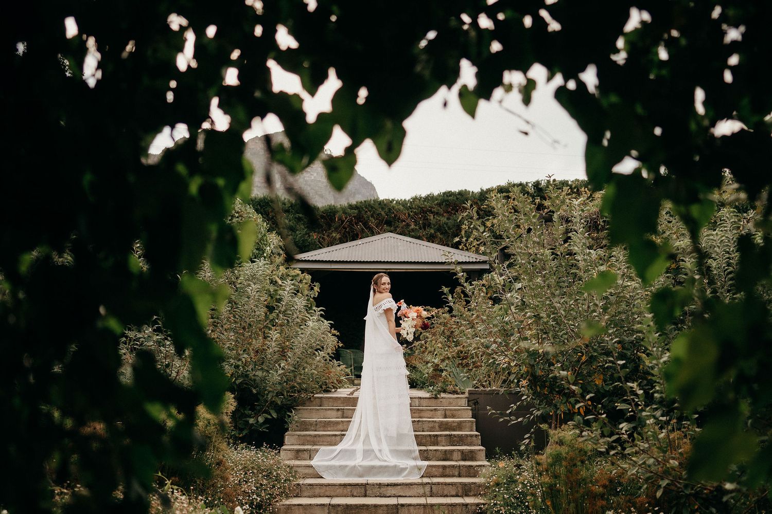 Figure in white dress with umbrella stands atop garden steps under elegant gazebo framed by lush greenery.